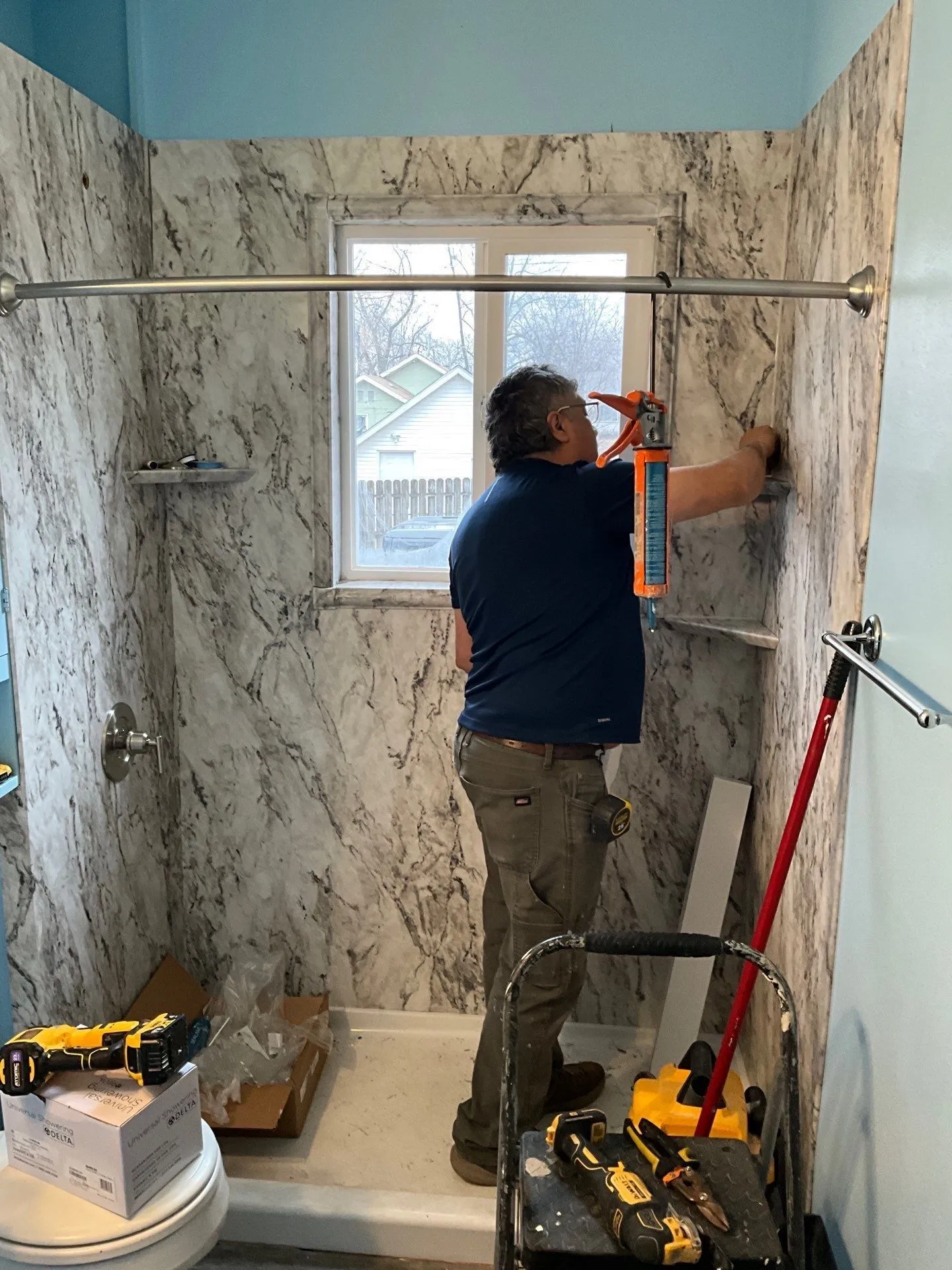 A Perfect Choice installer in a navy polo sealing the edge of a marble-look shower surround, with tools and packaging on the floor.
