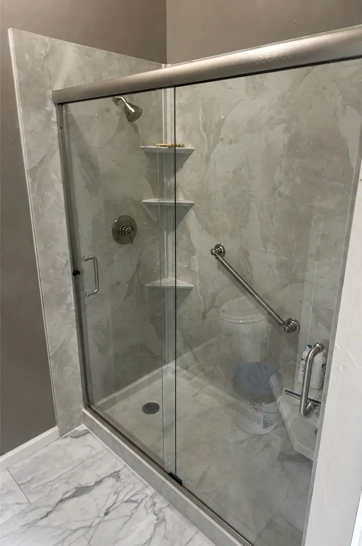 A finished grey marble-look walk-in shower with two grab bars, three corner shelves, a moulded corner seat and a low threshold.