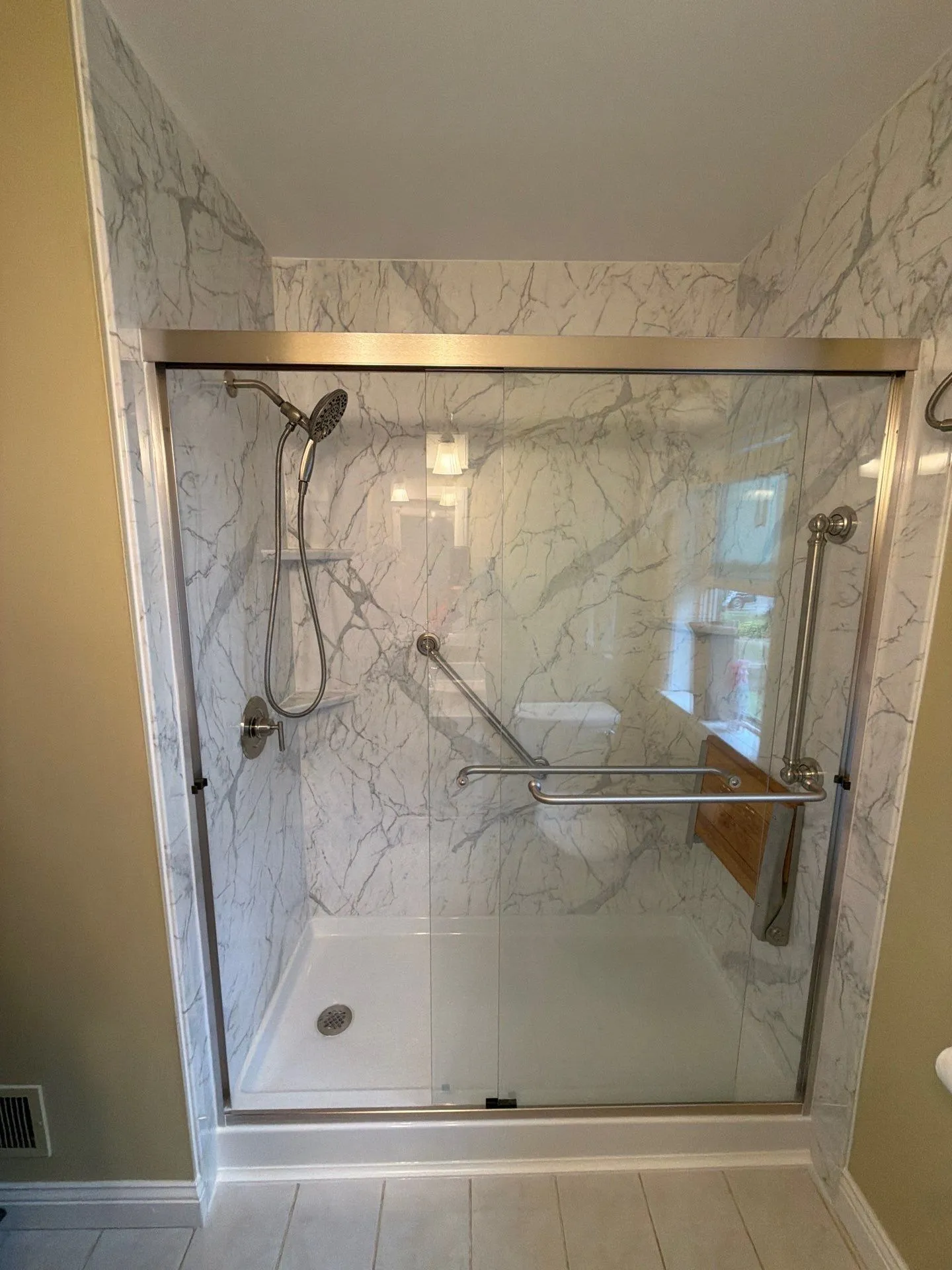 A finished walk-in shower with marble-look walls, a fold-down teak seat, two grab bars and a low threshold.